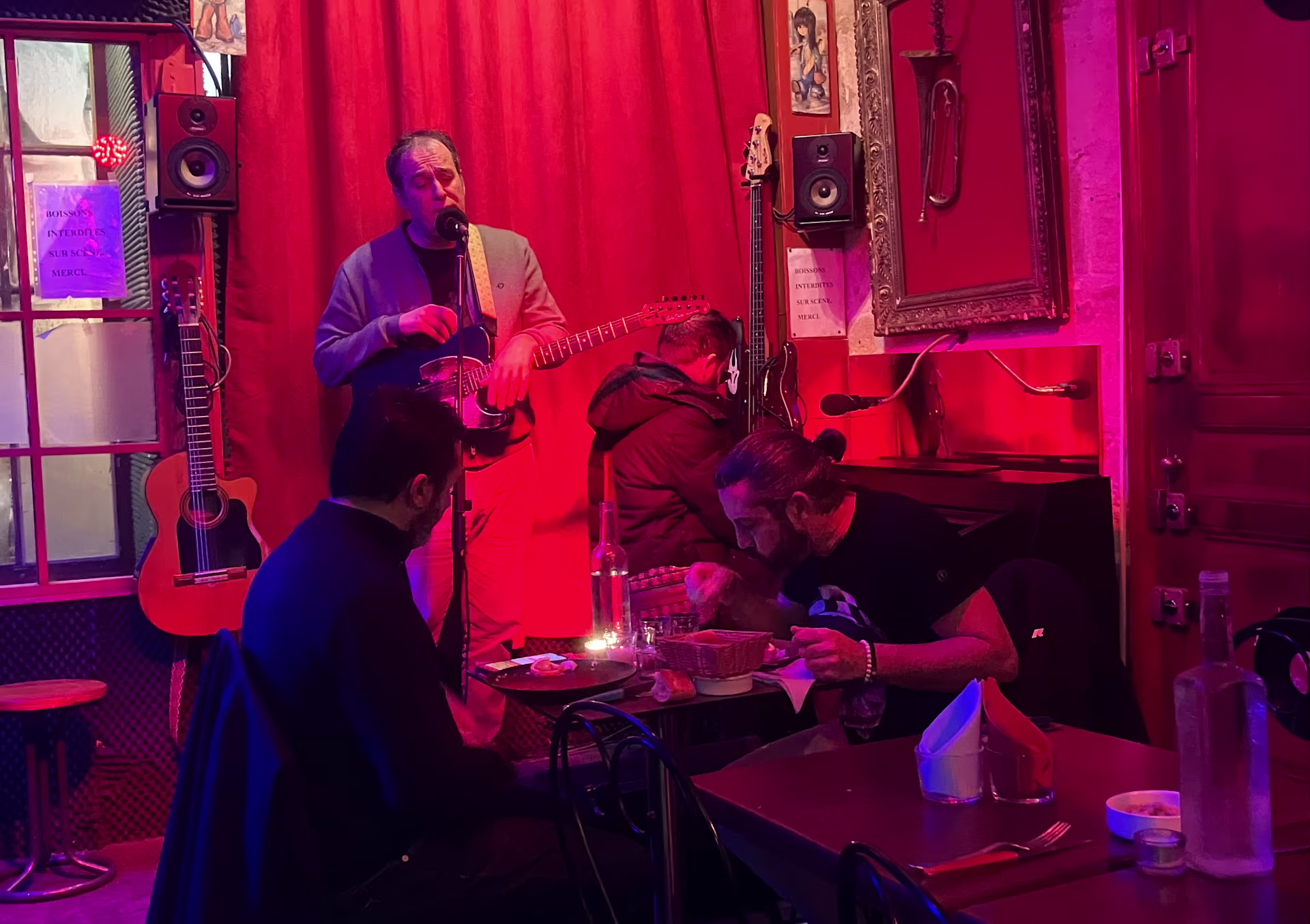 Int&eacute;rieur de restaurant anim&eacute; avec une sc&egrave;ne pour un musicien, murs d&eacute;cor&eacute;s d'&oelig;uvres encadr&eacute;es et &eacute;clairage tamis&eacute; rouge, cr&eacute;ant une atmosph&egrave;re chaleureuse.