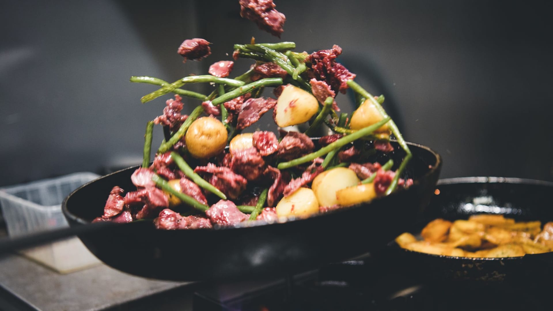 Sc&egrave;ne de cuisine dynamique avec des morceaux de viande marin&eacute;e, des pommes de terre dor&eacute;es et des haricots verts croquants, sugg&eacute;rant un processus de cuisson &eacute;nergique.