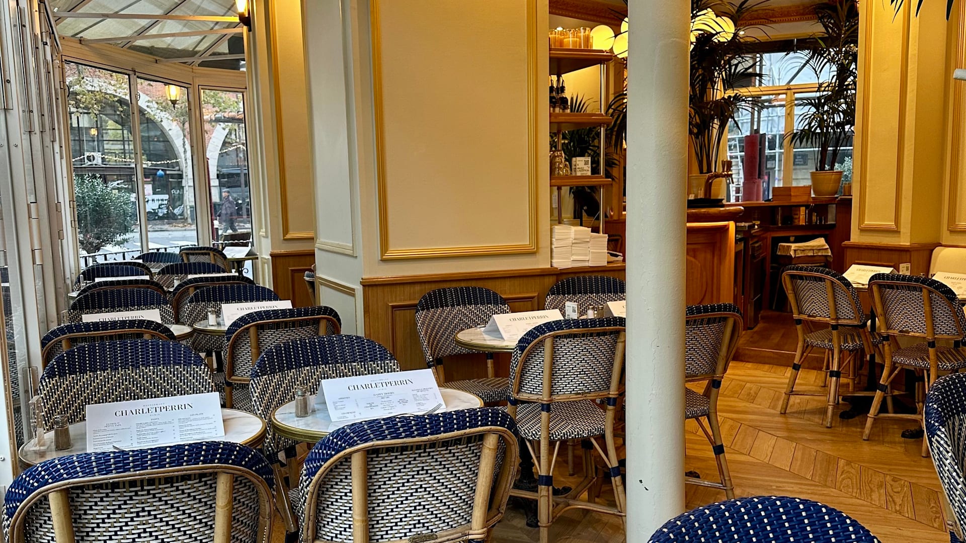 Int&eacute;rieur de restaurant chaleureux avec des tables petites entour&eacute;es de chaises en osier, murs clairs et accents en bois, baign&eacute; de lumi&egrave;re naturelle et agr&eacute;ment&eacute; de plantes en pot.