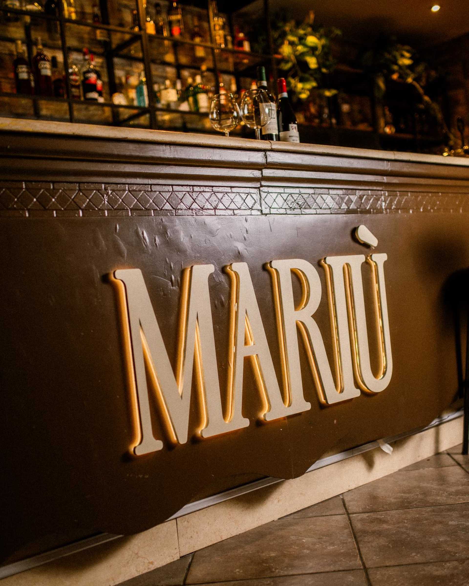 Area bar elegante con insegna 'MARIÙ', finiture in legno scuro e illuminazione raffinata, con scaffali pieni di diverse bottiglie.