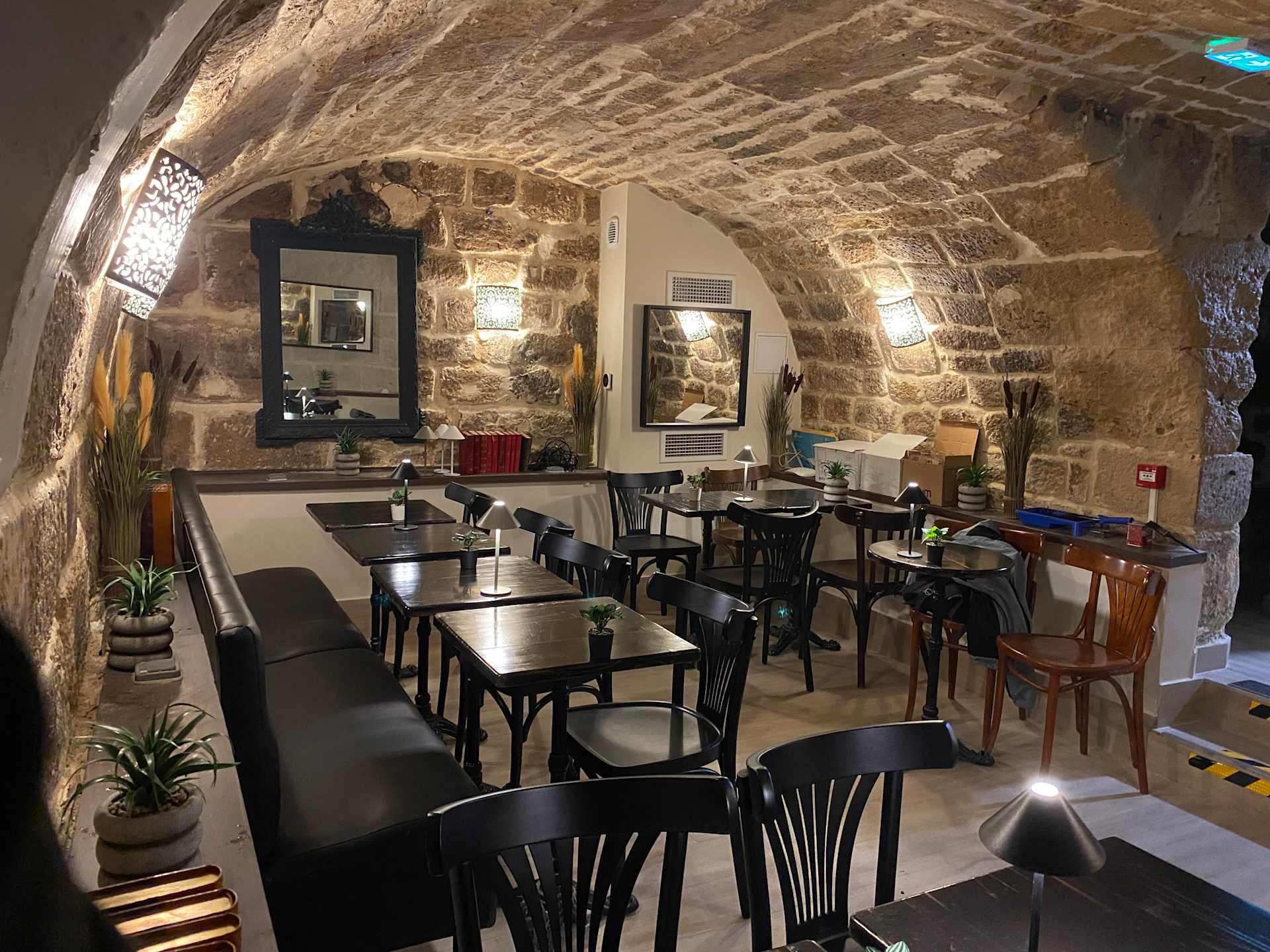 Int&eacute;rieur de restaurant charmant avec une arche en pierre rustique, &eacute;clairage chaleureux, tables et chaises en bois et noir, cr&eacute;ant une atmosph&egrave;re accueillante. Des plantes en pot ajoutent une touche de verdure, et un grand miroir refl&egrave;te l'ambiance conviviale. Design alliant &eacute;l&eacute;ments traditionnels et touche moderne pour un environnement de restauration confortable.