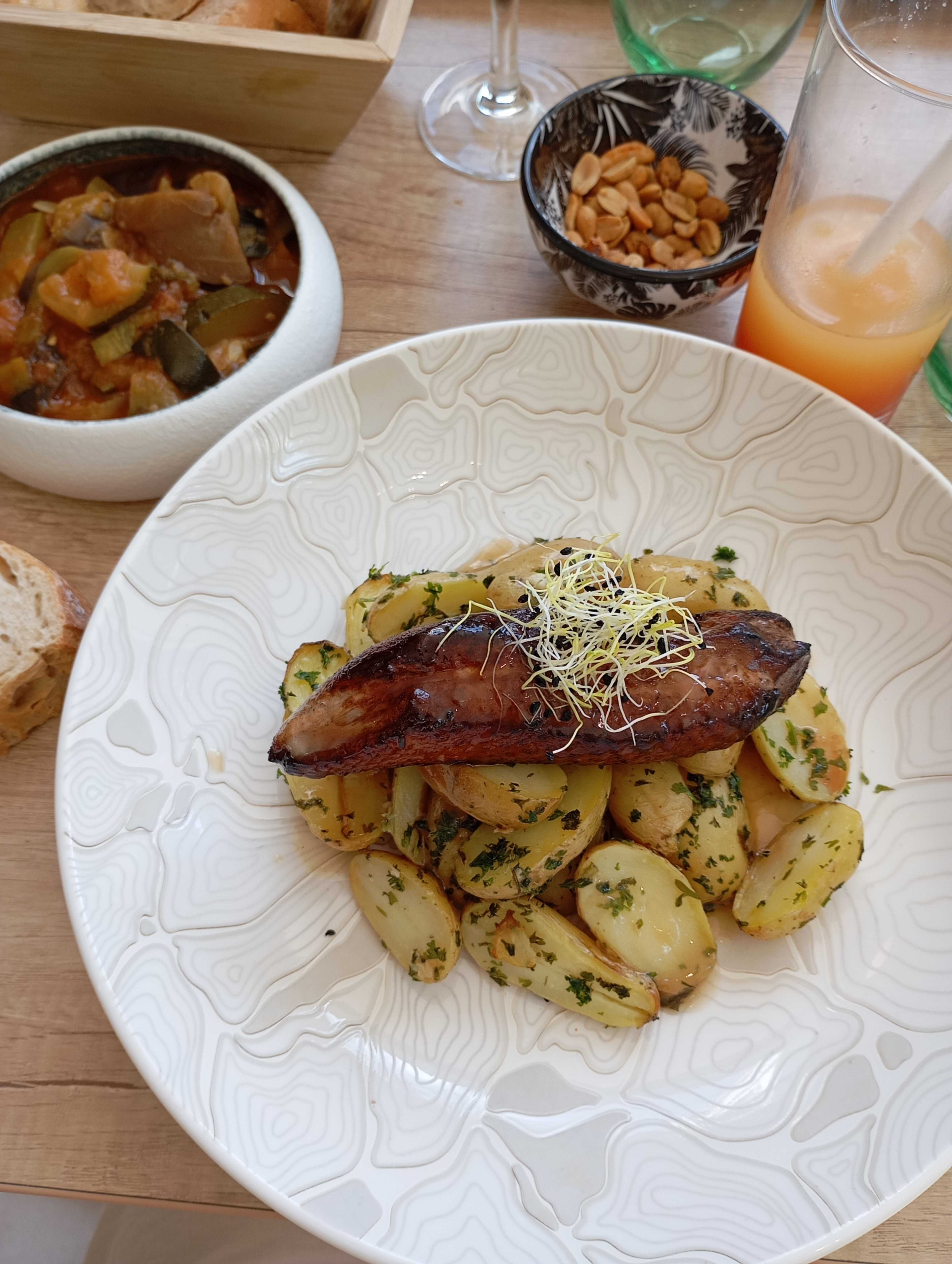Assiette de pommes de terre nouvelles rôties, assaisonnées et garnies d'herbes fraîches, accompagnées d'une viande grillée, probablement du porc ou du poulet, et d'un bol de ratatouille colorée avec des légumes variés, ainsi qu'un côté de noix croquantes.