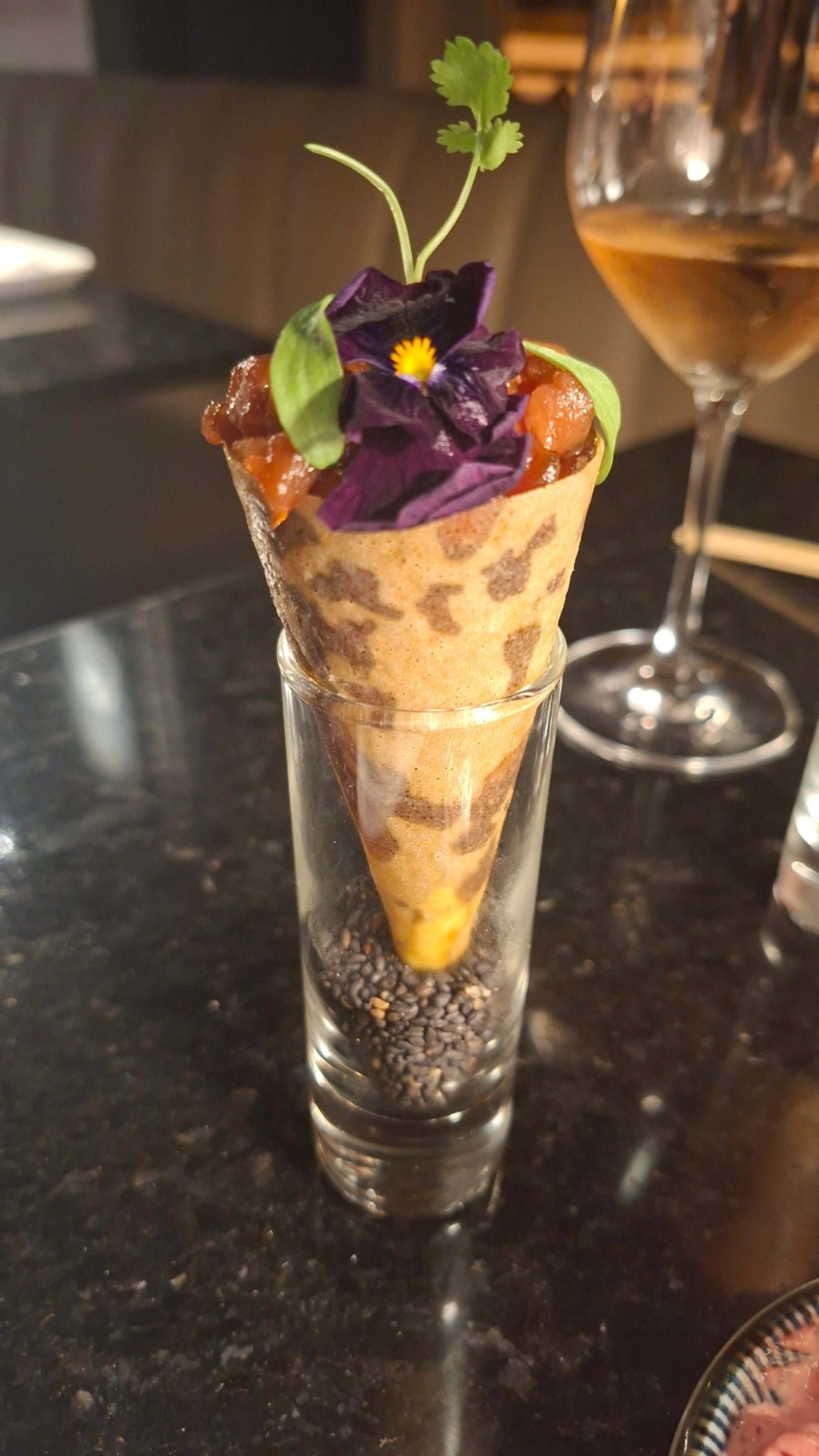 Prato único servido num recipiente com formato de cone com uma mistura saborosa coberta por um molho brilhante, guarnecido com uma flor comestível roxa e uma erva verde. A base do cone repousa em sementes de sésamo preto, realçando a apresentação.