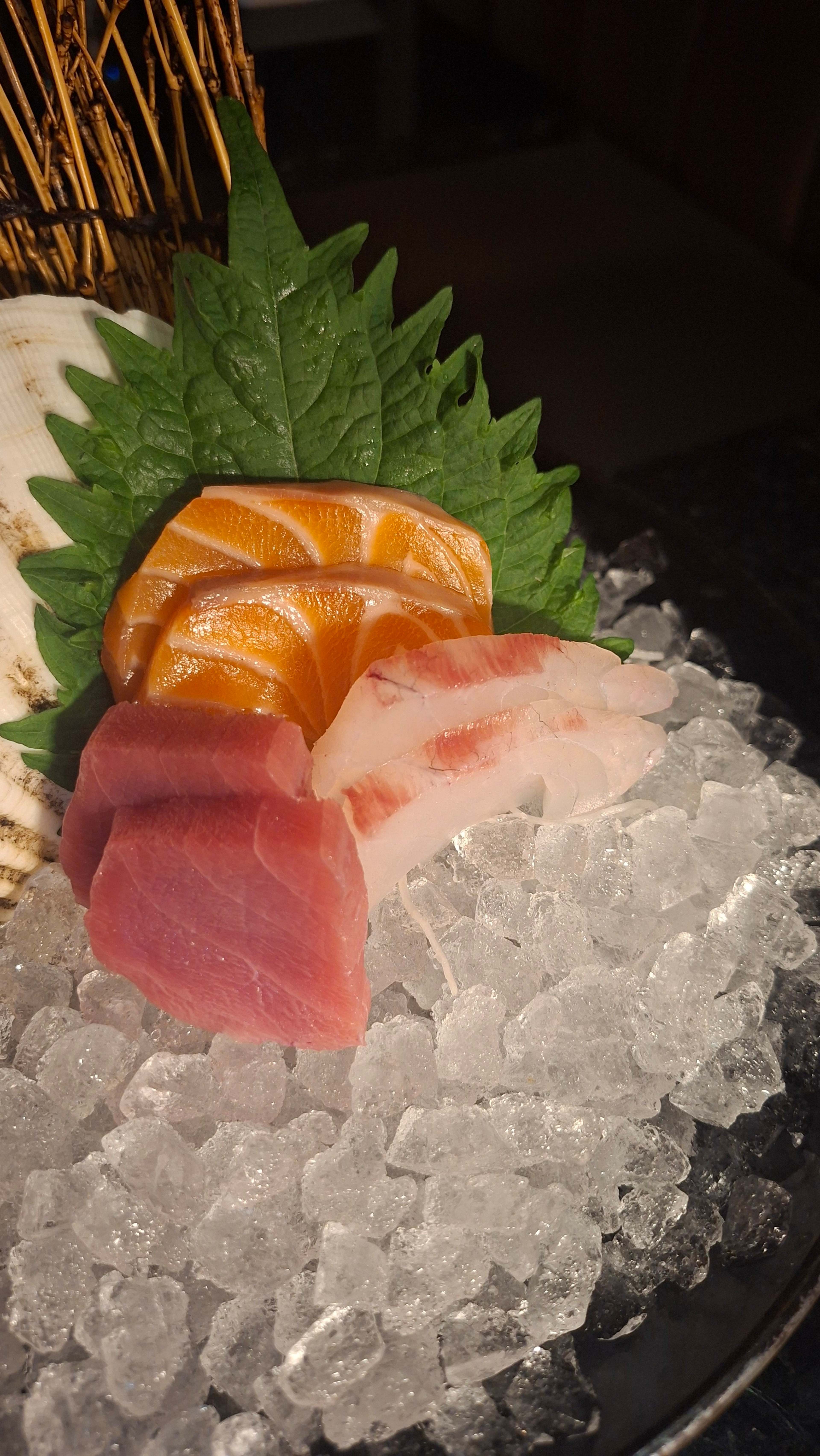 Prato de sashimi com três tipos de peixe: salmão laranja, atum rosa e um peixe branco, provavelmente pargo, sobre uma cama de gelo picado, guarnecido com folhas de shiso frescas.
