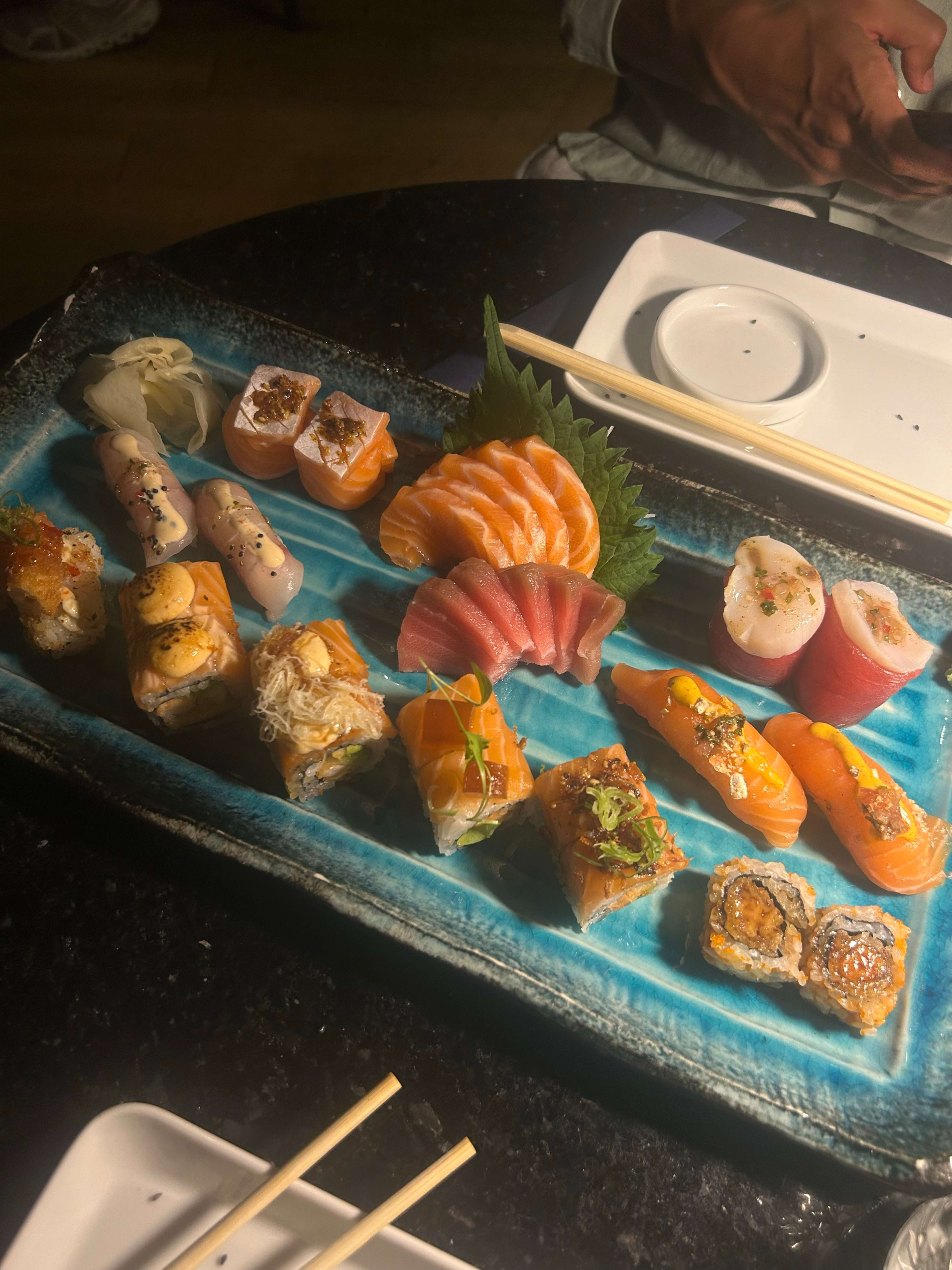 Um sortido de sushi disposto num prato azul, apresentando uma variedade de rolls e sashimi, incluindo sashimi de salmão cortado em forma de leque e rolls coloridos recheados com ingredientes como caranguejo, abacate e pepino, alguns com guarnições delicadas como sementes de sésamo e cebolinhas.