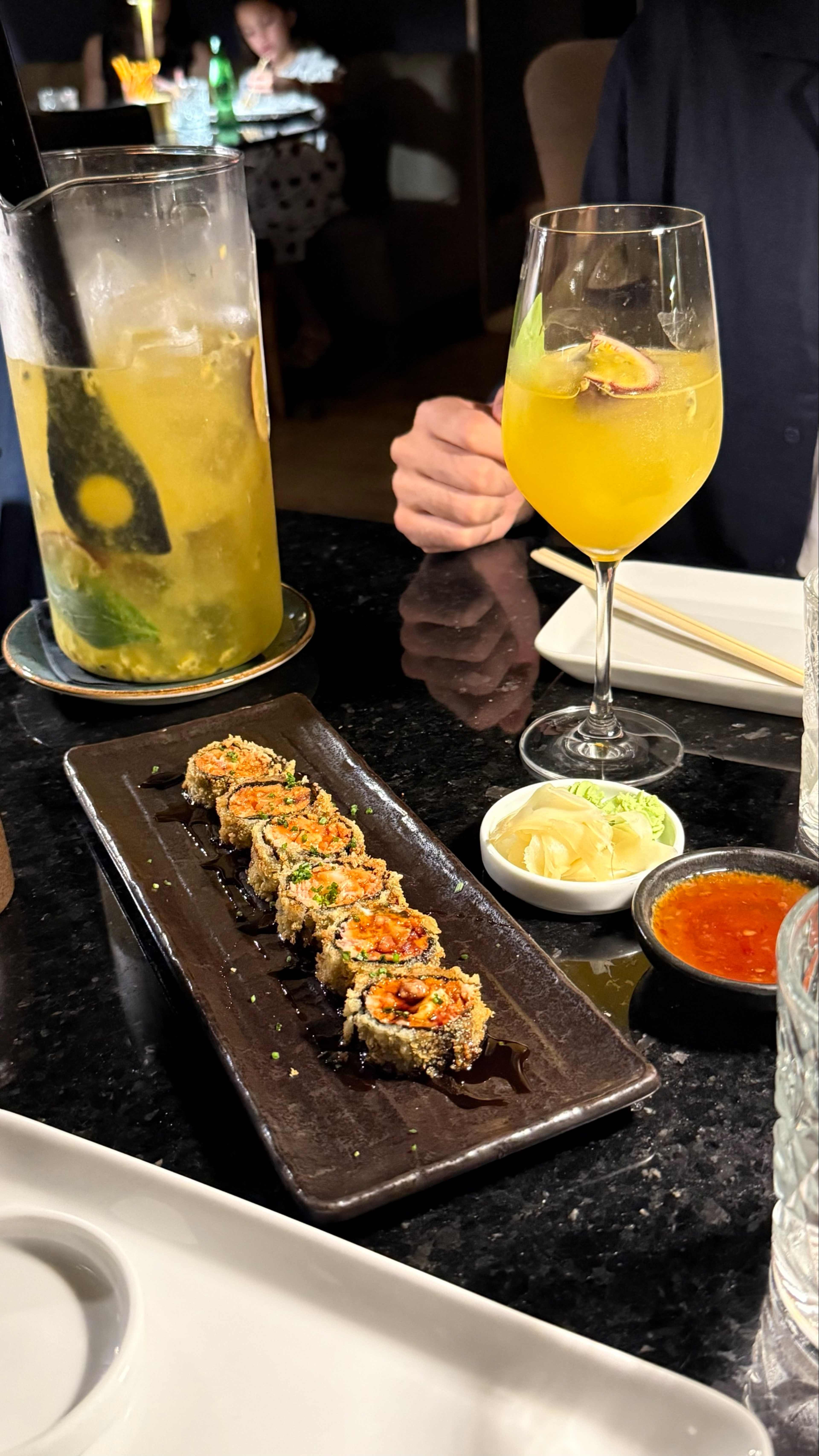 Prato de sushi com rolos variados, cobertos com uma textura crocante e guarnecidos com ervas picadas e um molho escuro. Acompanha gengibre em conserva e um molho picante.