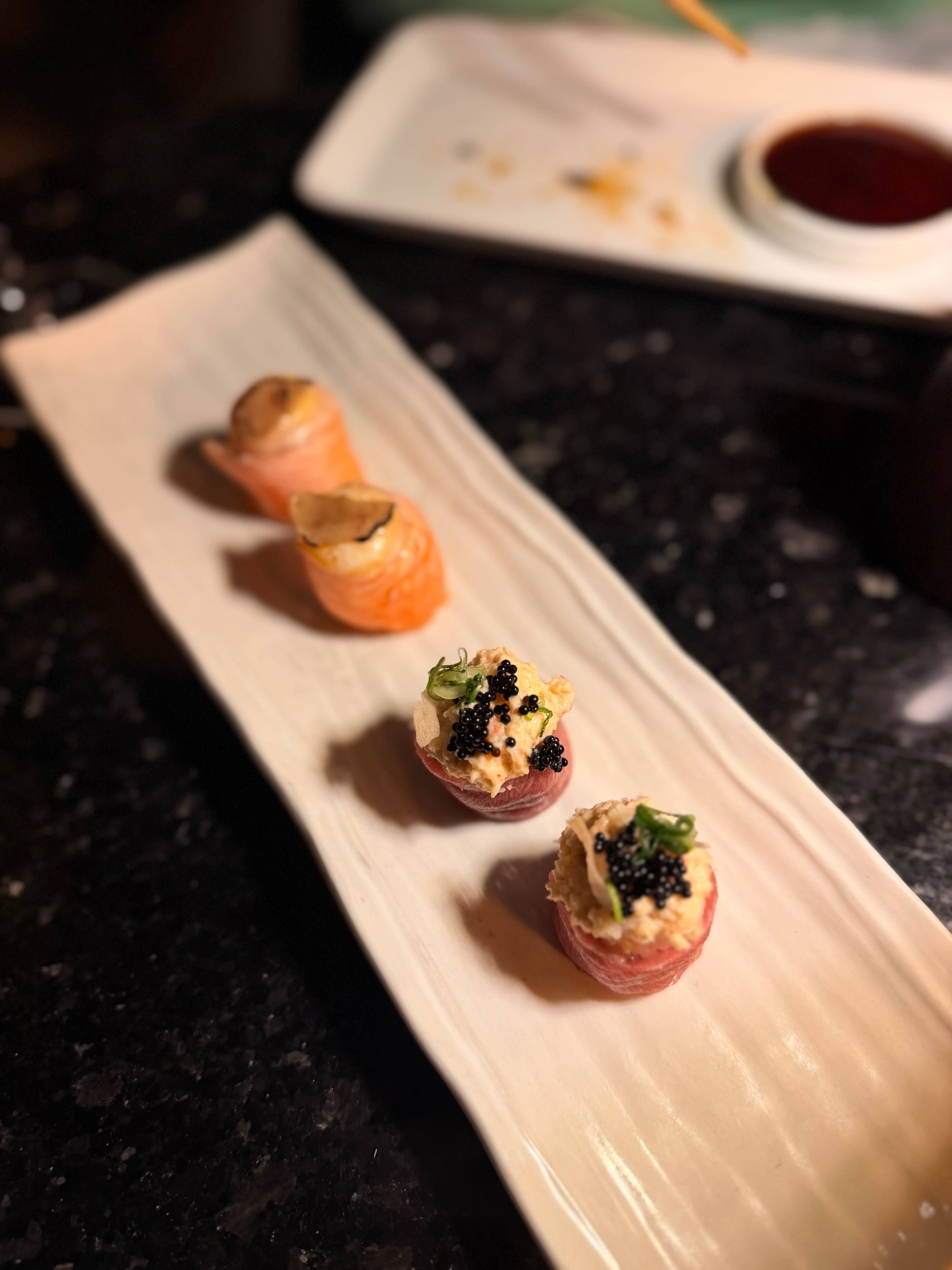 Apresentação delicada de sushi com quatro peças, sendo a primeira coberta com uma fatia de salmão e a segunda com um recheio cremoso, guarnecido com caviar negro e ervas finamente picadas. As cores suaves e convidativas destacam o salmão rosa em contraste com o caviar escuro.