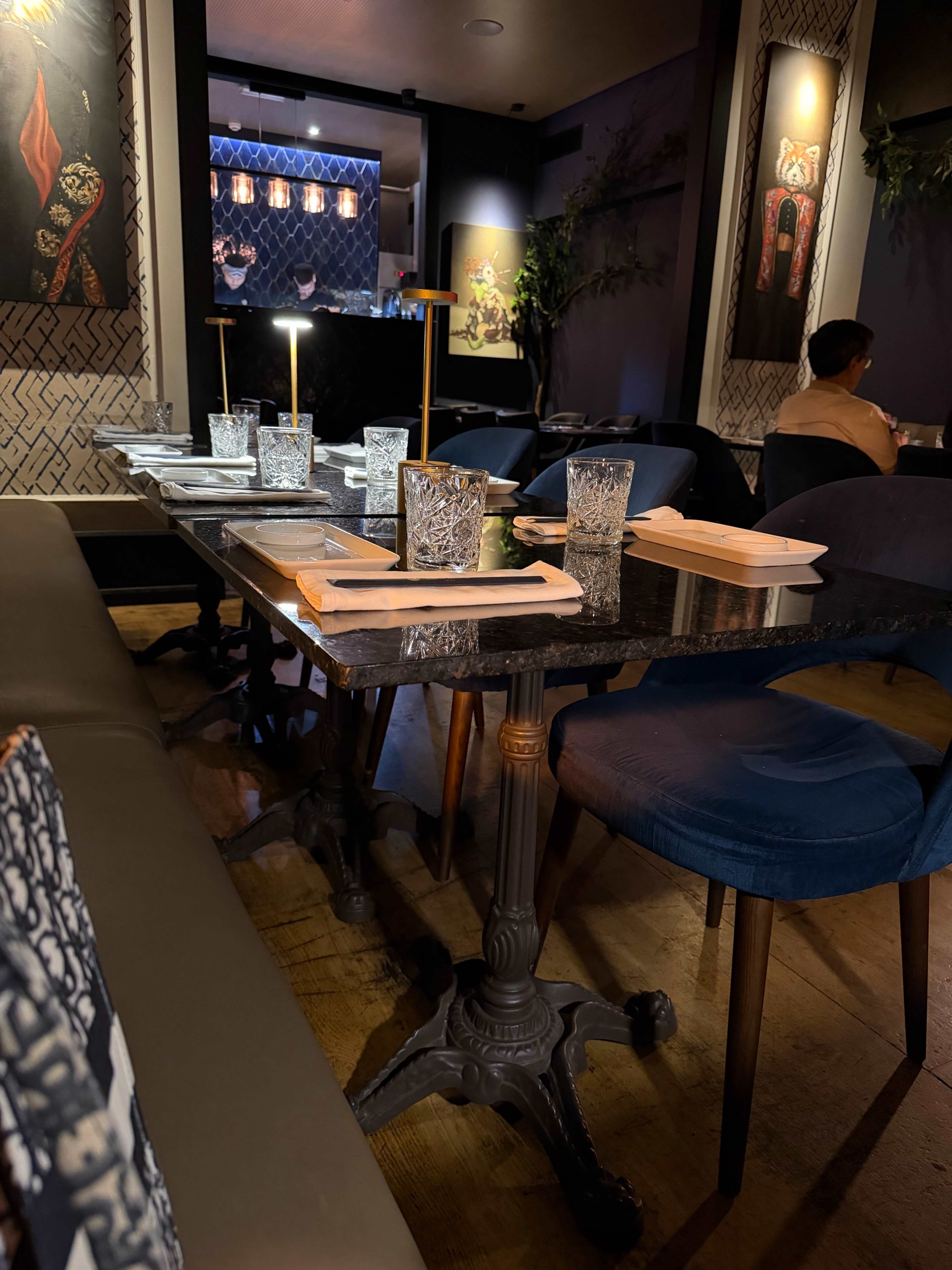 Interior de restaurante elegante com mesas de madeira escura e cadeiras azuis acolchoadas, iluminação suave e arte de parede decorativa, criando uma atmosfera acolhedora e sofisticada.