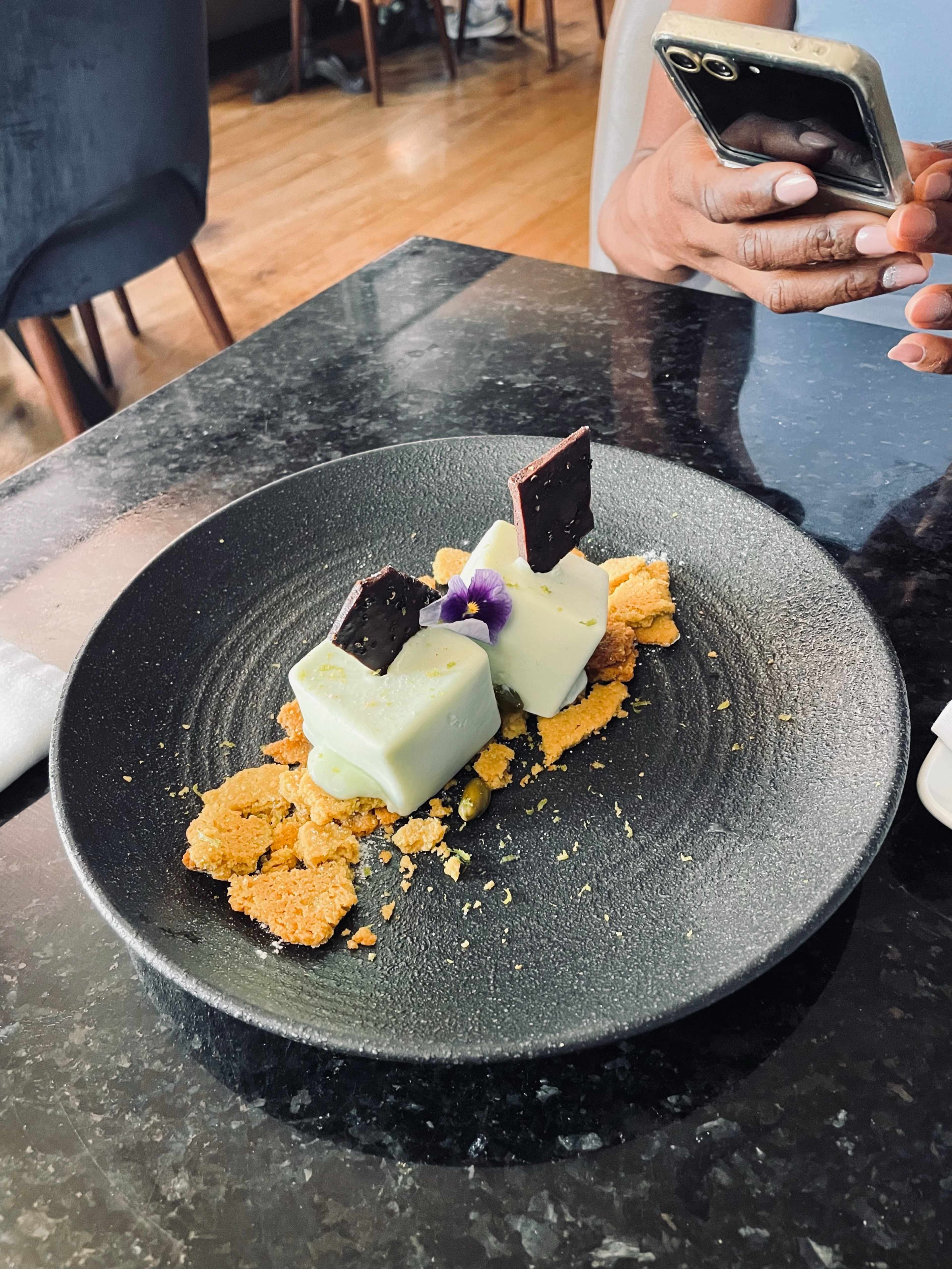Sobremesa delicada composta por dois blocos retangulares de mousse verde pálido, possivelmente aromatizada com matcha ou pistácio, guarnecida com quadrados de chocolate negro e uma pequena flor comestível, acompanhada de pedaços esfarelados de uma base crocante dourada.