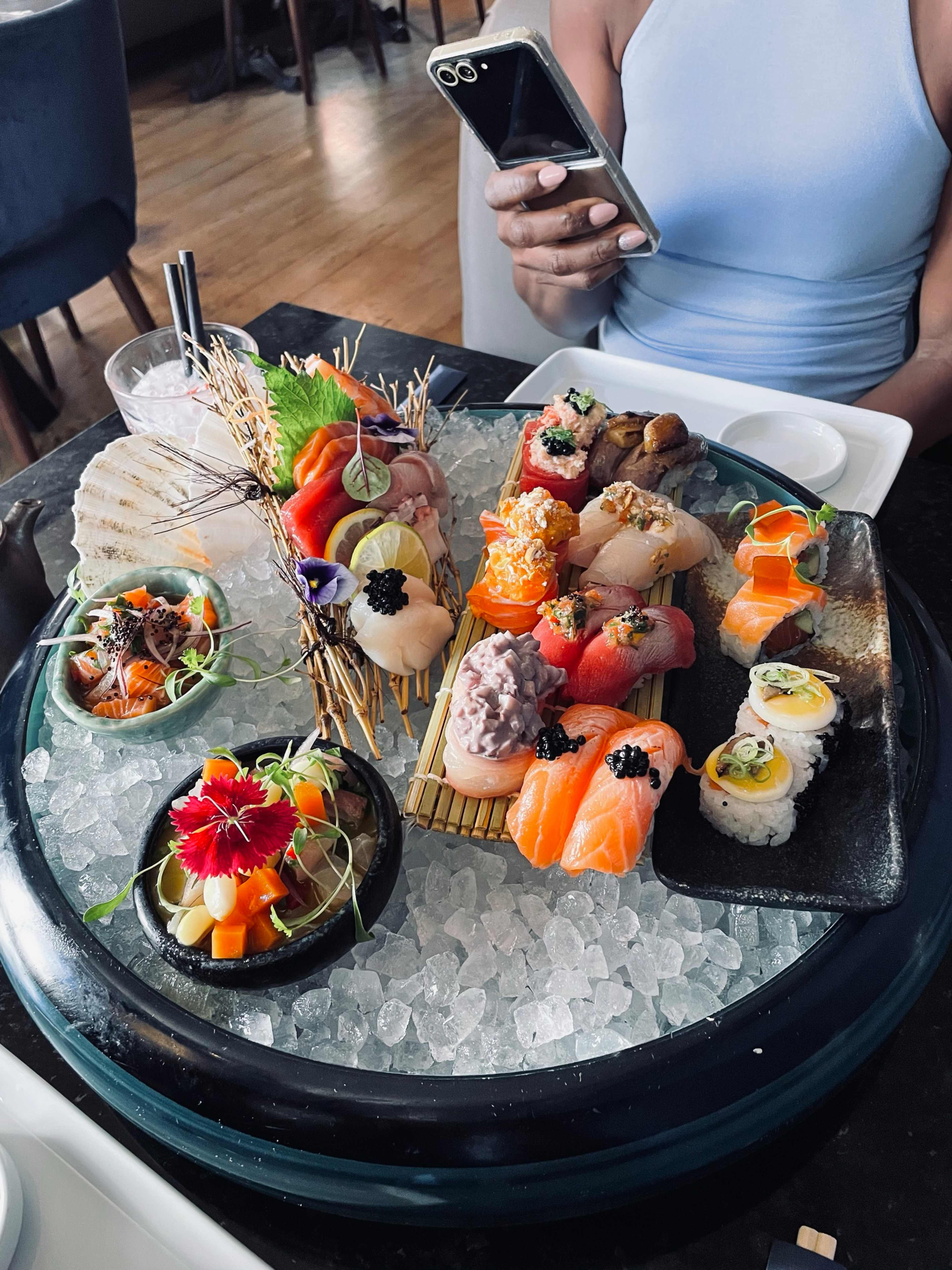 Prato elegante de sushi com uma variedade de sushi e sashimi, incluindo nigiri coloridos com peixe fresco, como salmão e atum, guarnecidos com caviar. Acompanha fatias vibrantes de legumes e ervas, além de sashimi de peixe fatiado. Elementos decorativos de flores comestíveis realçam a apresentação, servidos sobre uma cama de gelo.