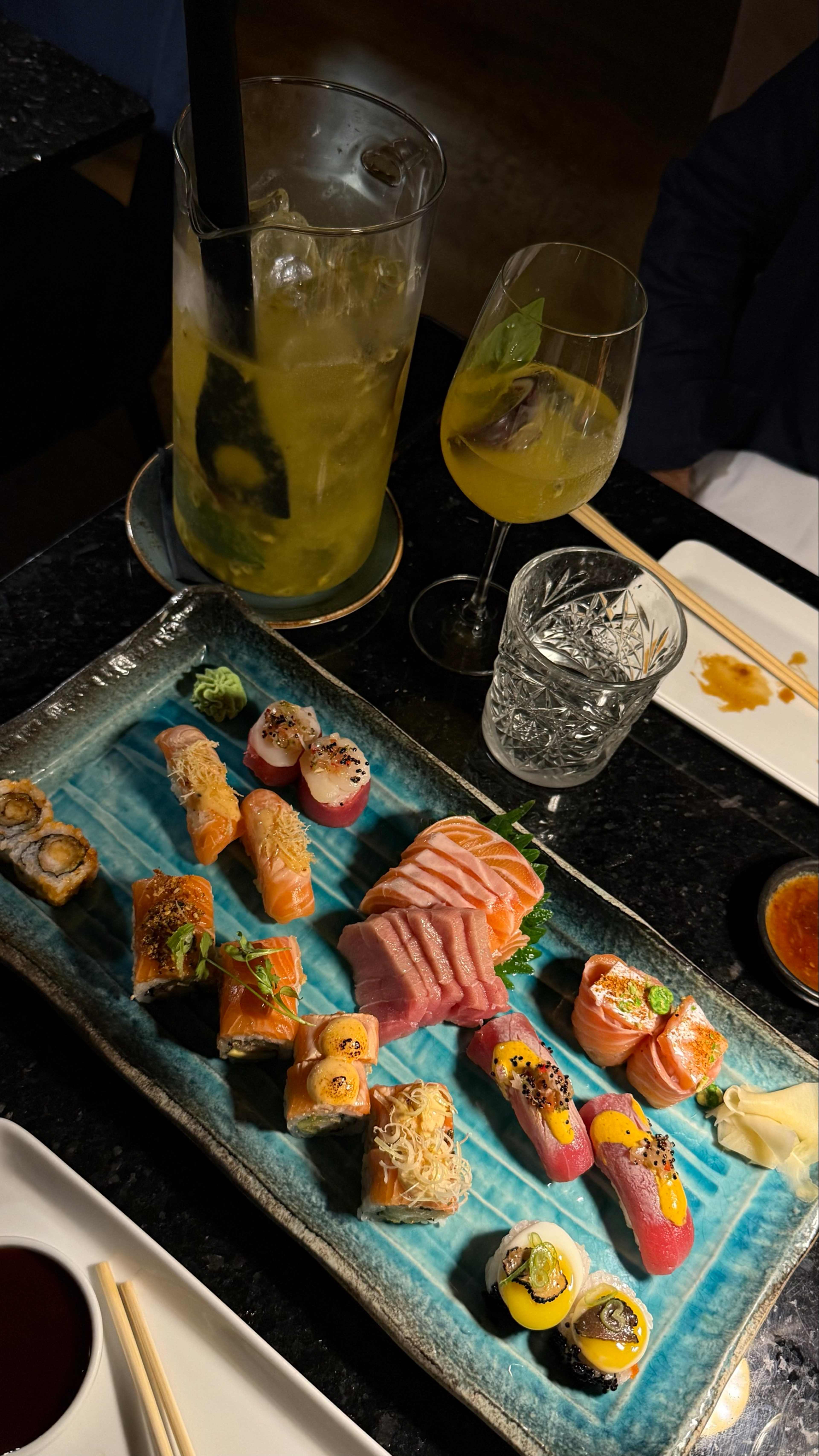 Prato de sushi bem apresentado com nigiri de salmão, atum e peixe-gato, além de rolos, incluindo um rolo de tempura crocante e um rolo arco-íris. Guarnições de wasabi e gengibre em conserva realçam a apresentação.
