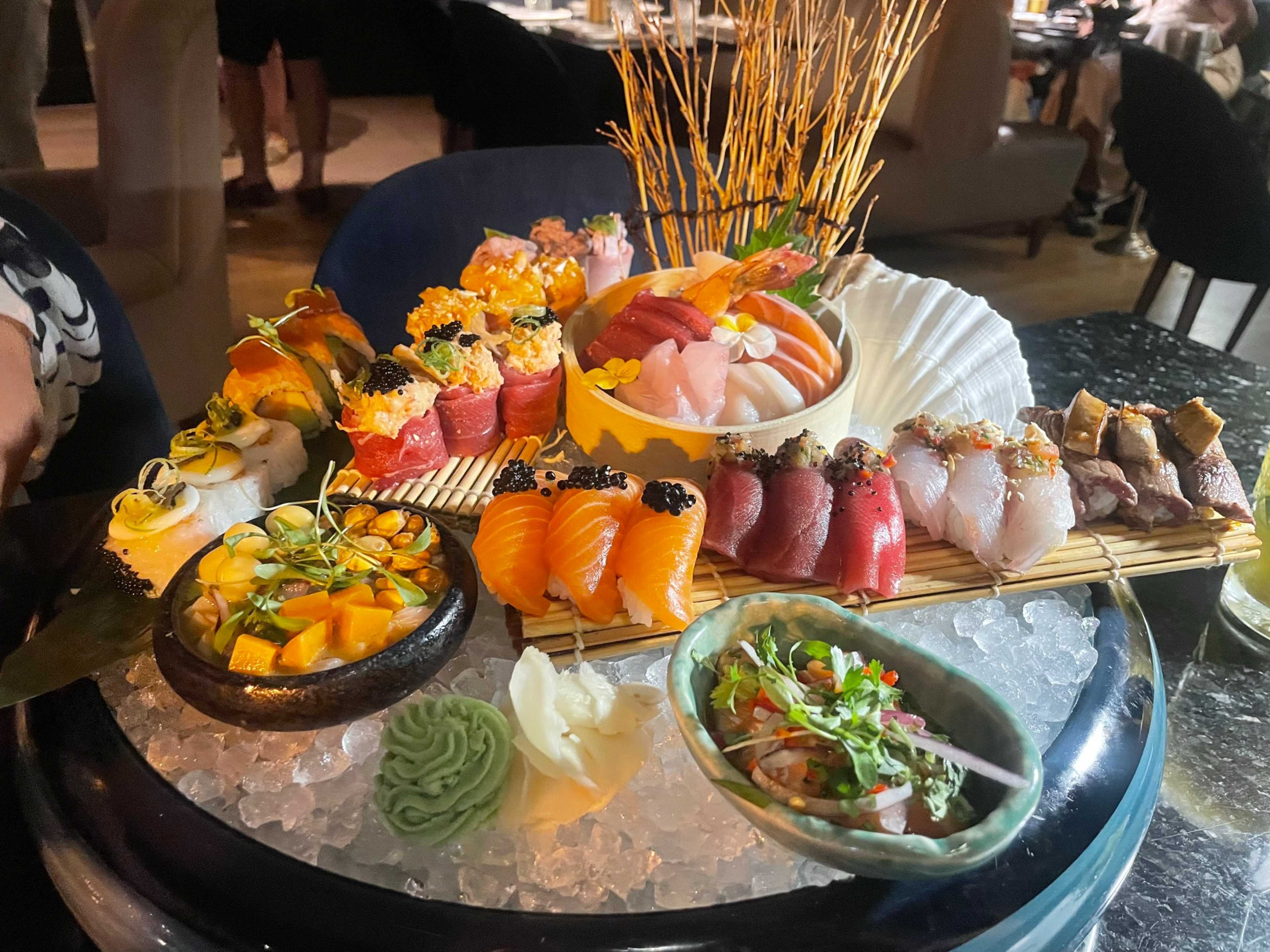 Prato elaborado de sushi com uma variedade de nigiri e sashimi, incluindo fatias coloridas de salmão, atum e peixe-gato, guarnecidas com caviar e microvegetais. Acompanha uma salada refrescante de manga e pepino.