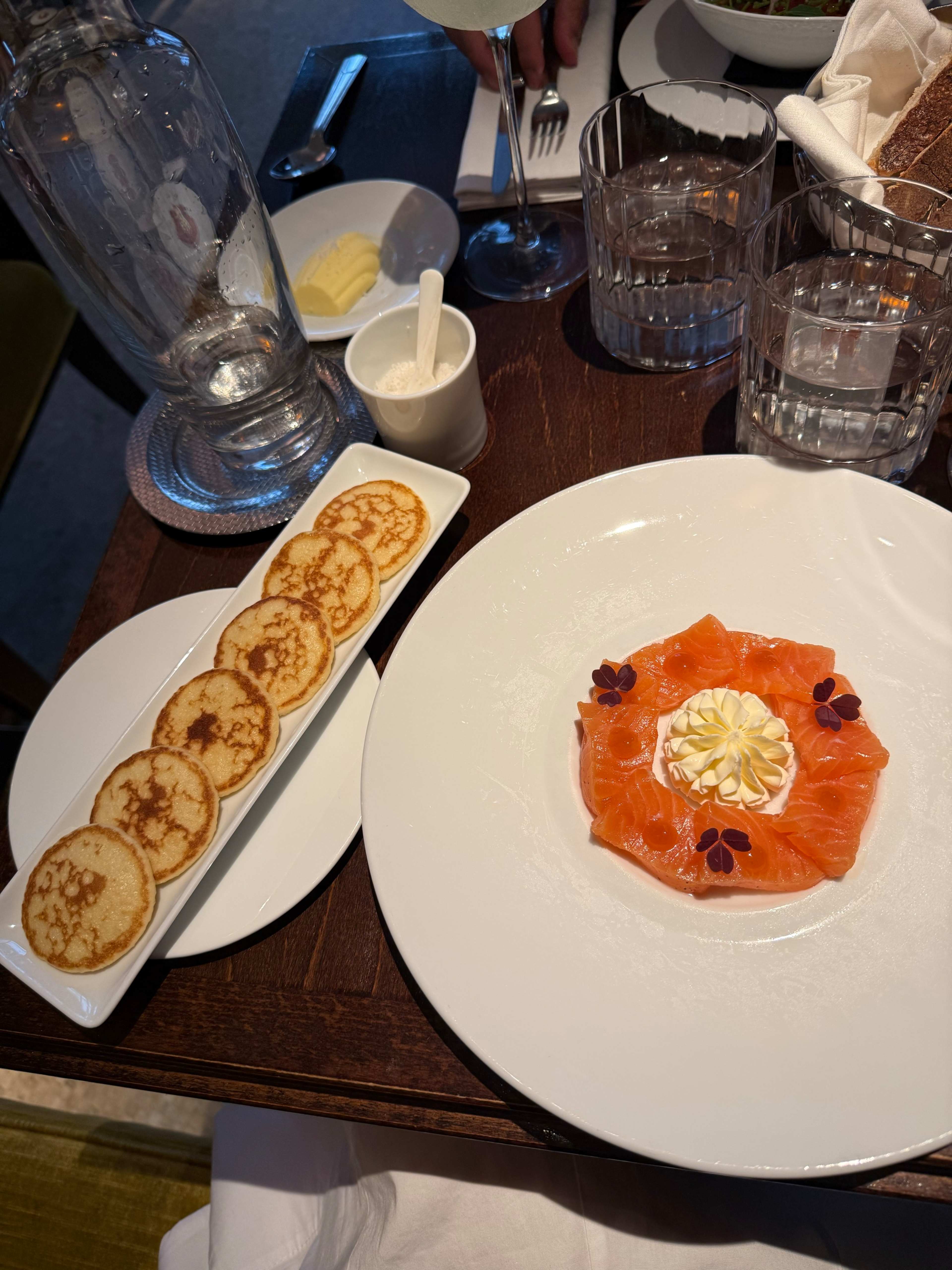 Plat délicatement présenté avec du saumon fumé tranché finement, garni de crème et de fleurs comestibles, accompagné de pancakes ronds légèrement grillés.