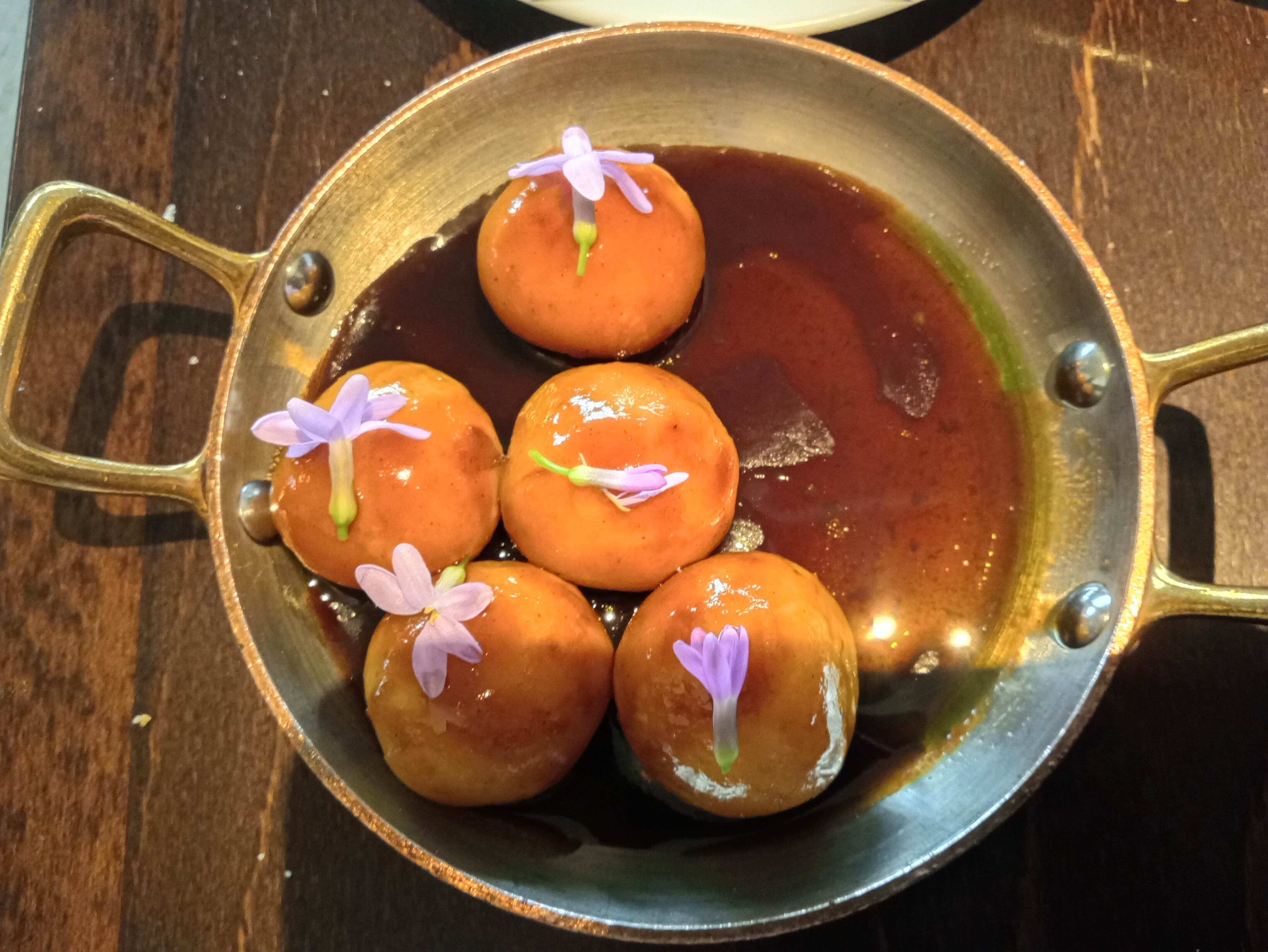 Plat délicat composé de six boulettes dorées, probablement à base de pâte sucrée, servies dans un sirop foncé et garnies de petites fleurs violettes.