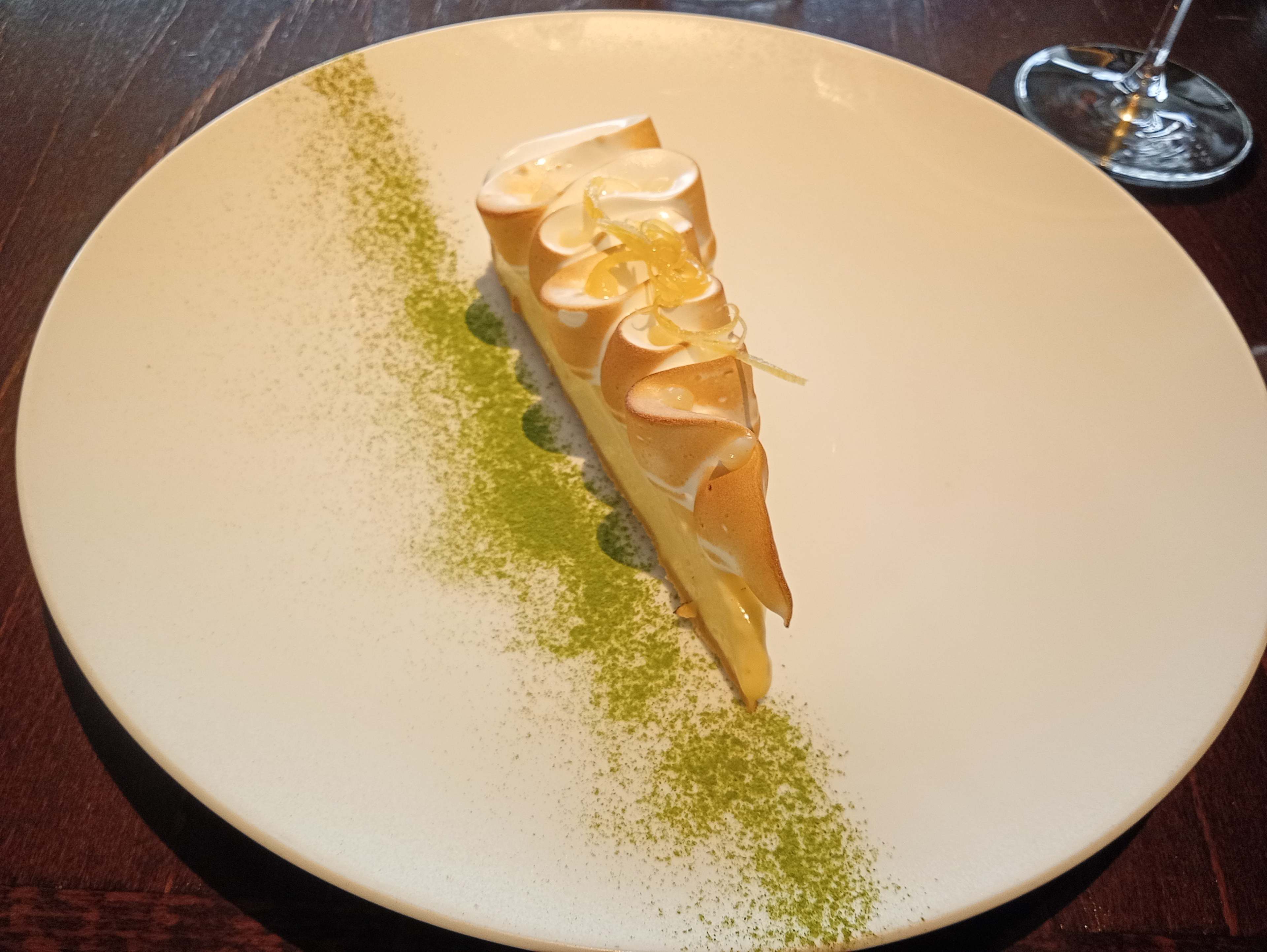 Une part de tarte au citron élégamment présentée, avec une garniture crémeuse et lisse, surmontée de meringue dorée et de zestes de citron. Un saupoudrage de poudre de matcha vert ajoute un contraste saisissant.