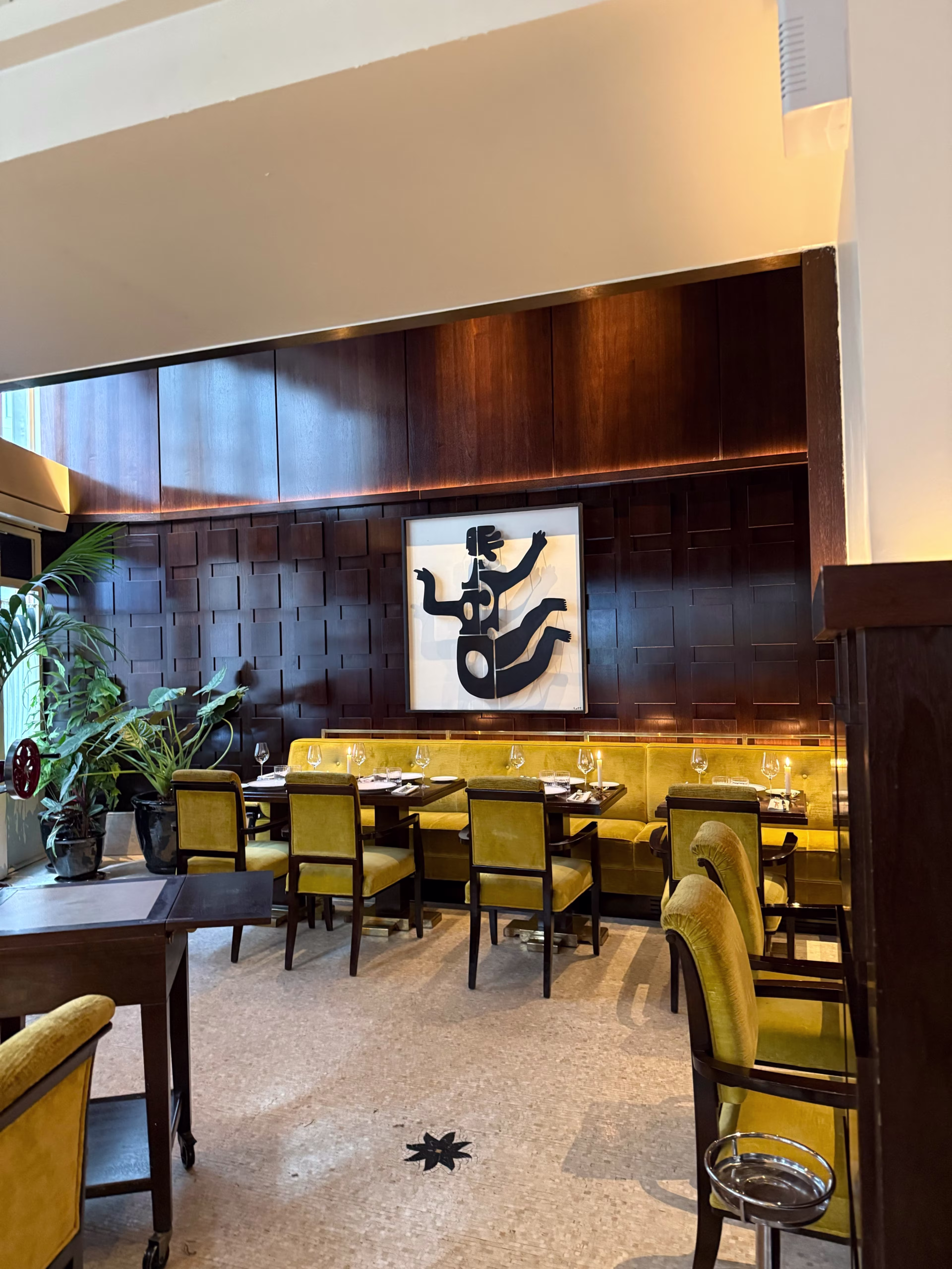 Intérieur de restaurant élégant avec des panneaux en bois foncé et des sièges jaunes doux, décoré d'une grande œuvre d'art abstraite. Ambiance chaleureuse et sophistiquée, avec des tables élégamment dressées et des plantes en pot.