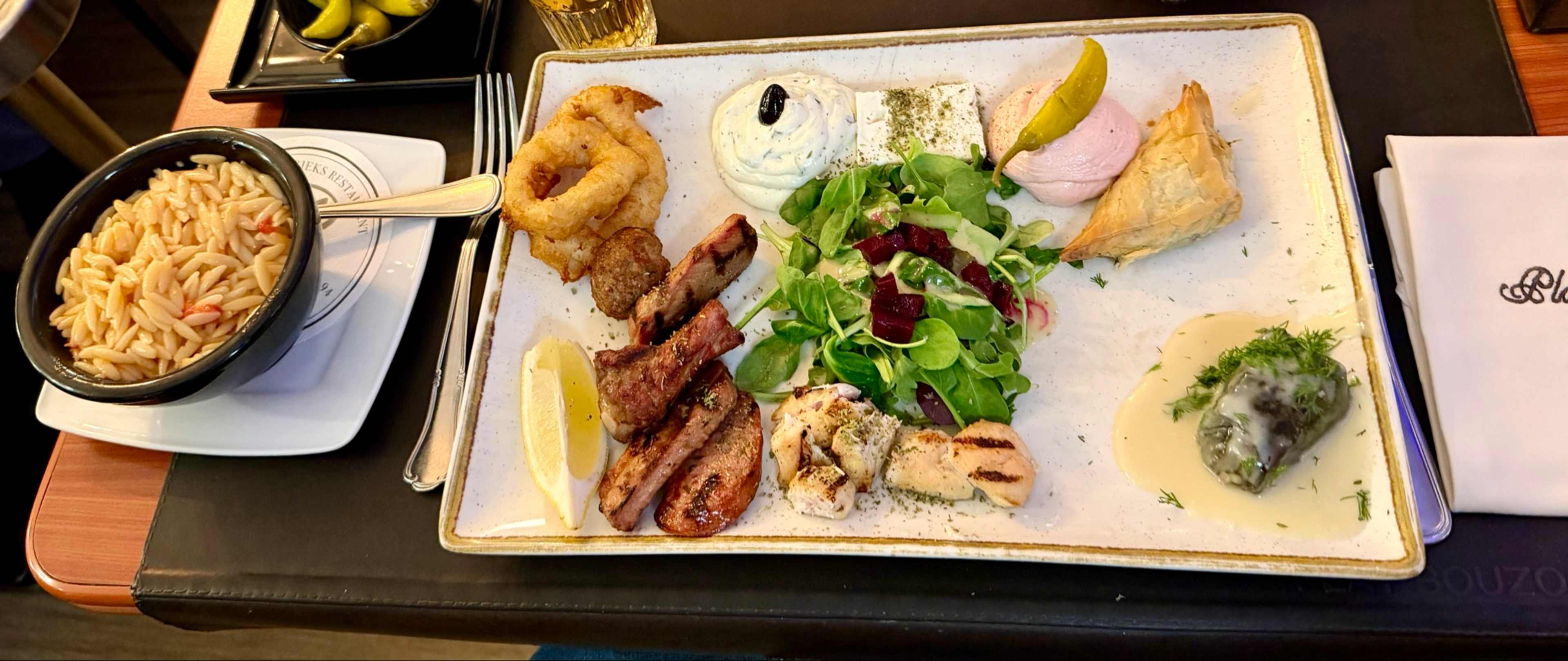 Mooi gepresenteerd plateau met gegrilde kip en rundvlees, gegarneerd met een schijfje citroen, vergezeld van knapperige calamaresringen en een schepje romige saus. Een frisse salade met gemengde groenten en cranberry's voegt kleur toe. Daarnaast een bladerdeeggebakje, mogelijk gevuld, en een klein portie groene kruiden, vermoedelijk een spinazie- of kruidenkroket. Een bijgerecht van orzo-pasta in een aparte kom maakt de diverse selectie compleet.