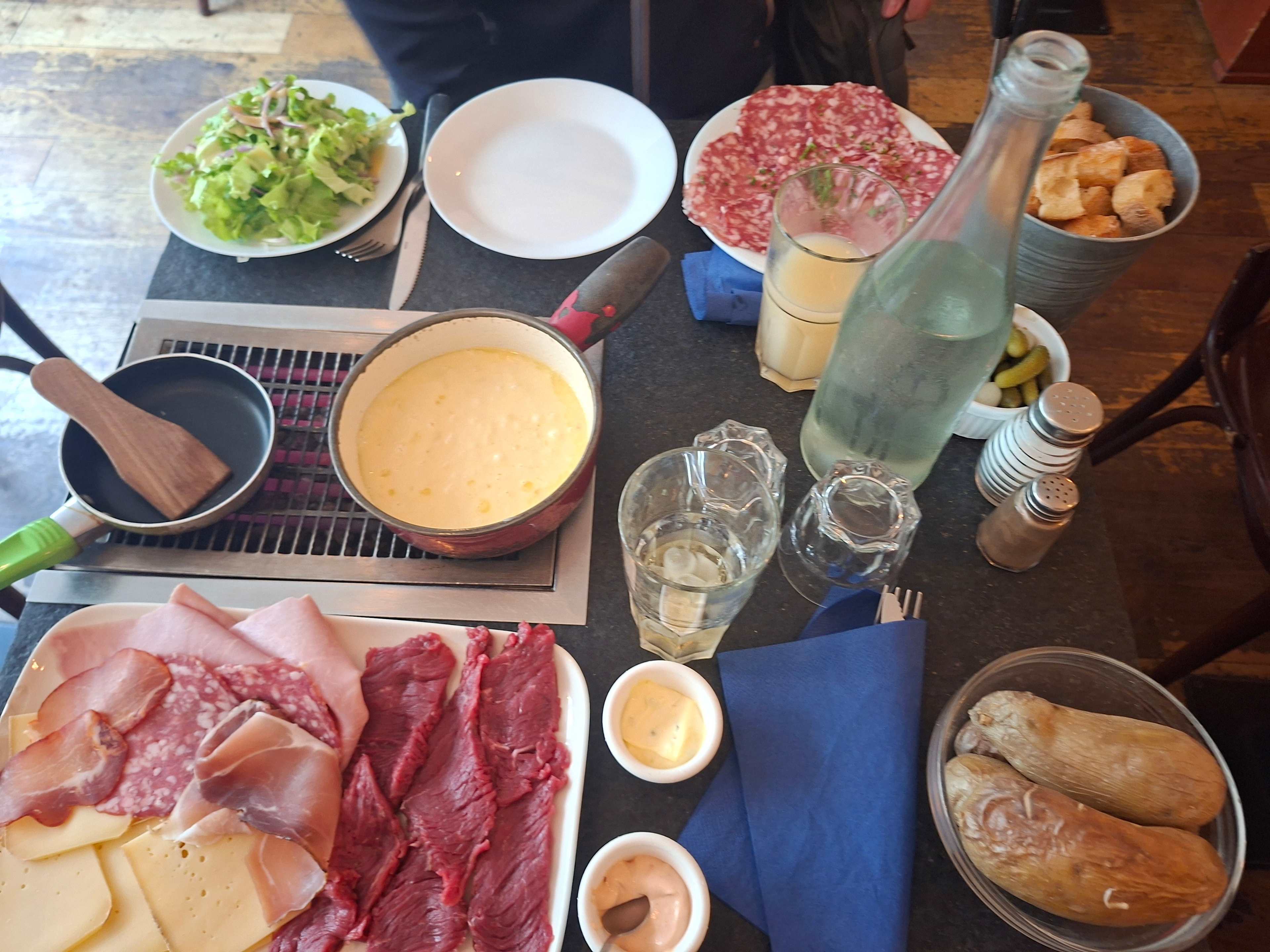 Sélection d'ingrédients pour un repas partagé, avec un pot de fromage fondu au centre, accompagné d'une petite poêle et d'une spatule en bois. Entouré de viandes, y compris du salami et du jambon tranchés, ainsi que de légumes frais en salade. Un panier de petits pains ajoute une touche rustique, tandis que les condiments et les boissons sont disposés de manière soignée sur la table, créant une atmosphère conviviale.