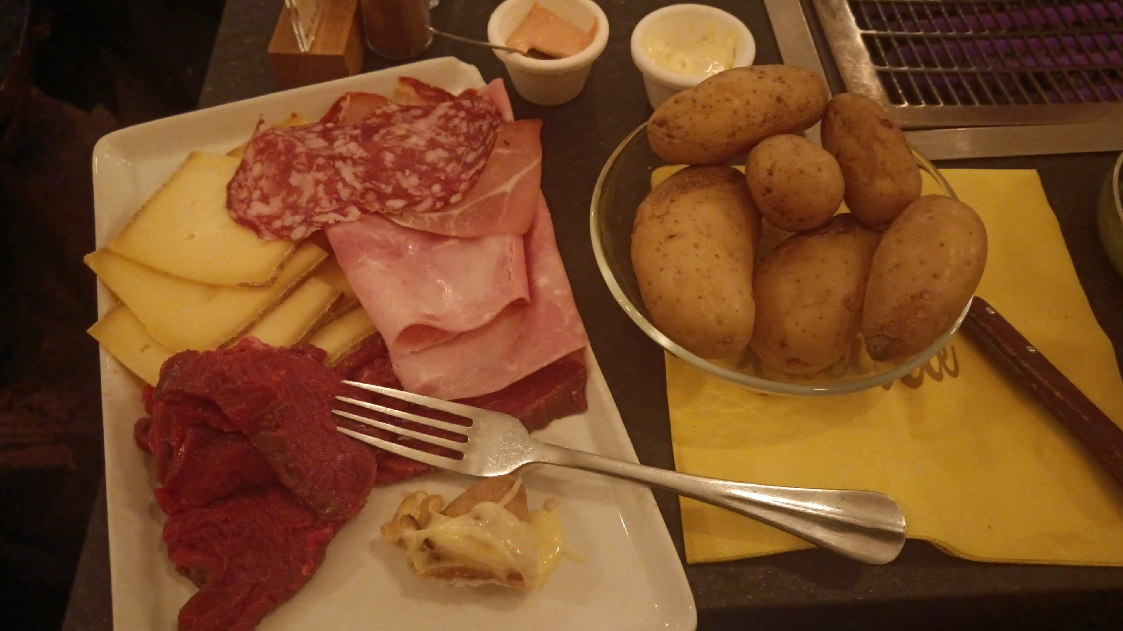 Plateau généreux avec un assortiment de charcuteries, comprenant des tranches de salami, de jambon et une variété de fromages, accompagné de petites coupelles de condiments et de pommes de terre bouillies.