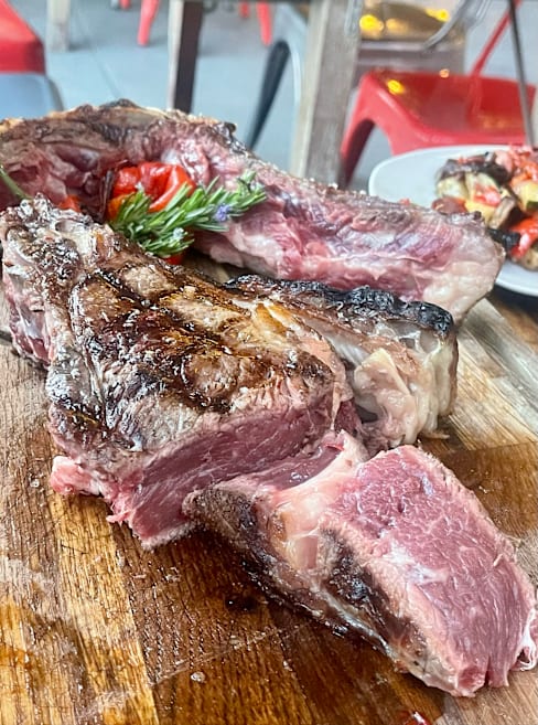 Carne a la parrilla, probablemente un ribeye, con textura marmoleada y acompañada de tomates asados y hierbas frescas.