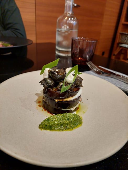 Plat en couches avec des tranches d'aubergine surmontées d'escargots grillés, accompagnés d'un pesto vert vibrant et de sauce crémeuse, le tout joliment présenté avec des oignons verts en garniture.