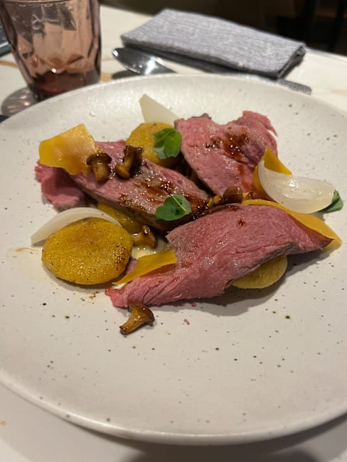 Plat élégamment présenté avec des tranches de viande tendre, accompagné d'un mélange de légumes jaunes et blancs, dont des morceaux grillés, et parsemé de champignons sautés, le tout garni d'herbes fraîches.