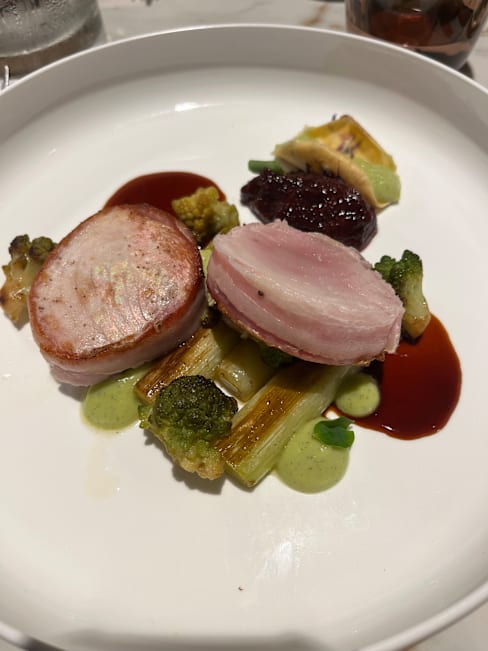 Plat joliment présenté avec deux tranches de porc tendre, l'une avec une croûte croustillante et l'autre avec un intérieur juteux. Accompagné d'un mélange de légumes, dont du brocoli grillé, et d'une sauce verte vibrante. Un ajout de compote de baies sucrée et un filet de sauce savoureuse complètent le tout.