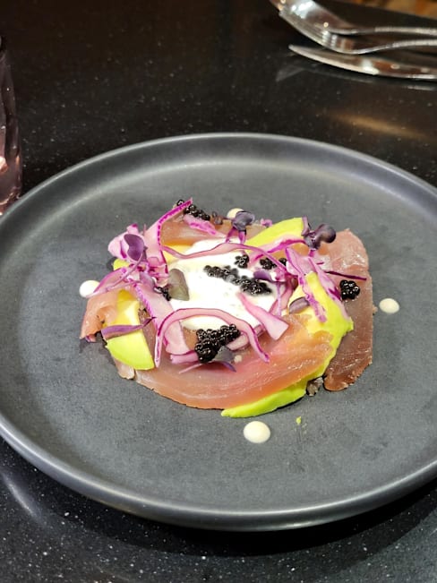 Plat joliment présenté avec des tranches de poisson, probablement thon, superposées avec de l'avocat et surmontées d'une sauce crémeuse, garnies de chou rouge vibrant et de perles de caviar.