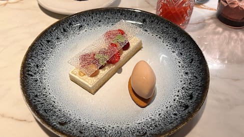 Dessert délicat composé d'une couche rectangulaire de cheesecake crémeux, garni de fraises fraîches et d'une feuille de sucre décorative, accompagné d'une boule de glace vanille sur un biscuit.