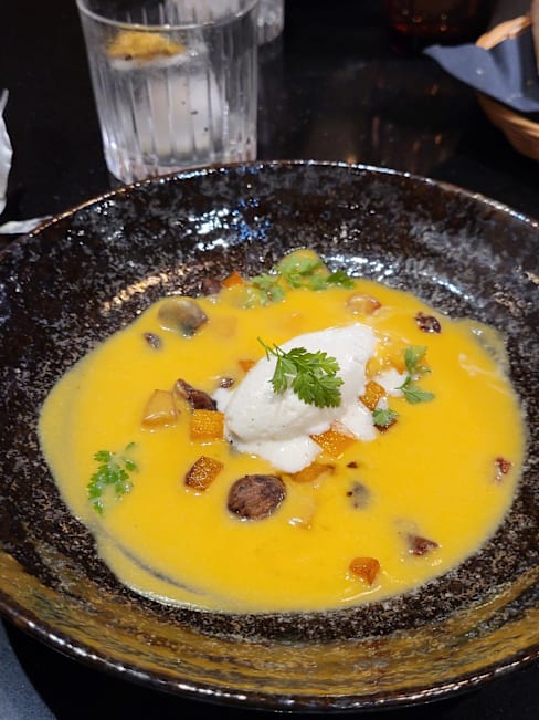 Soupe crémeuse à base de courge, agrémentée de morceaux de champignons grillés et de légumes en dés, surmontée d'une cuillerée de crème blanche et de fines herbes.