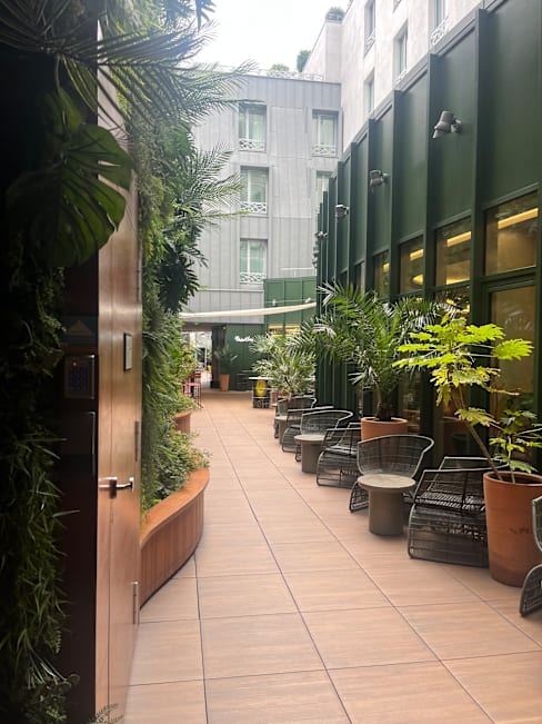Espace de restauration extérieur serein avec un chemin bordé de verdure luxuriante et de plantes en pot, entouré de sièges modernes et de bancs en bois, créant une atmosphère rafraîchissante et accueillante.
