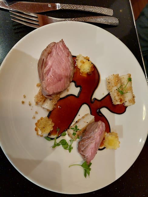 Plat composé de deux morceaux de viande tendre, probablement d'agneau ou de bœuf, entourés de croûtons dorés et d'une sauce épaisse, accompagnés de carrés crémeux de purée ou de mousse et garnis d'herbes fraîches.