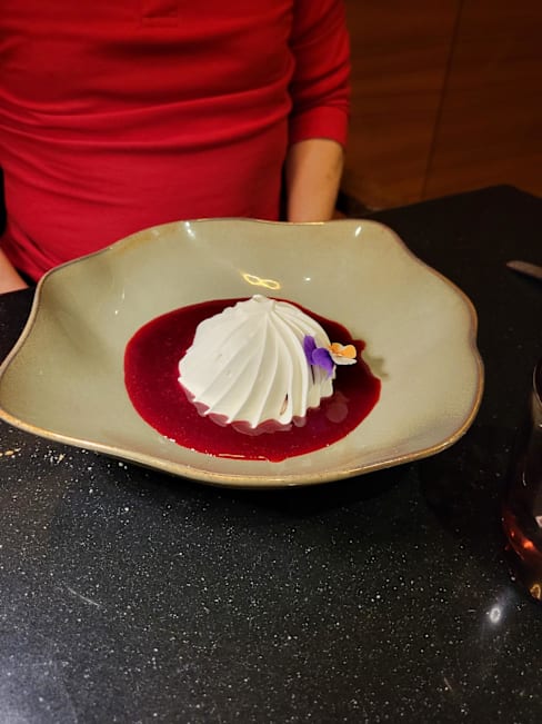 Dessert délicat avec une mousse élégamment façonnée, probablement à base de chocolat blanc ou de crème, sur un coulis de baies rouges brillant, garni d'une petite fleur comestible.