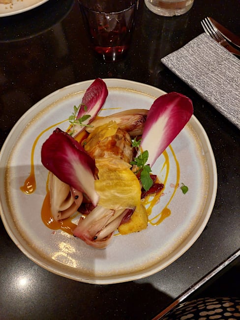 Plat élégamment présenté avec des morceaux tendres de viande, probablement du poulet, accompagnés d'un assortiment de légumes. La viande est nappée d'une sauce épaisse, rehaussant sa couleur dorée. Des tranches de radicchio grillé entourent la viande, ajoutant une touche de rouge profond et une légère amertume. Des garnitures délicates de légumes jaunes et blancs, peut-être marinés, offrent un contraste rafraîchissant. Un filet de sauce décore artistiquement l'assiette, tandis que de petites pousses ajoutent une note de fraîcheur à la présentation.