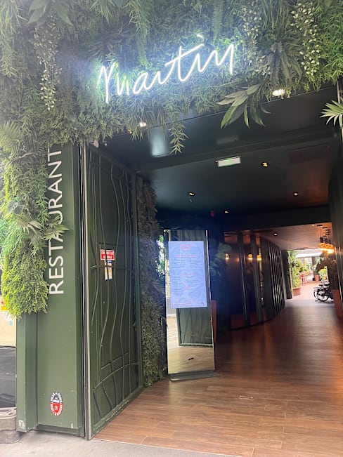 Entrée moderne d'un restaurant ornée de verdure, avec une façade vert foncé et un panneau lumineux indiquant 'Restaurant', un grand miroir et un éclairage chaleureux. Le sol en bois apporte une touche de chaleur au design contemporain.