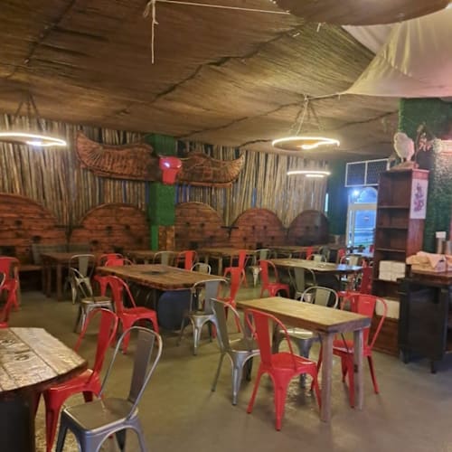 Ambiente rústico y casual con paredes de madera y techo de paja, asientos de sillas metálicas rojas y plateadas junto a mesas de madera, creando un espacio acogedor y bien iluminado.
