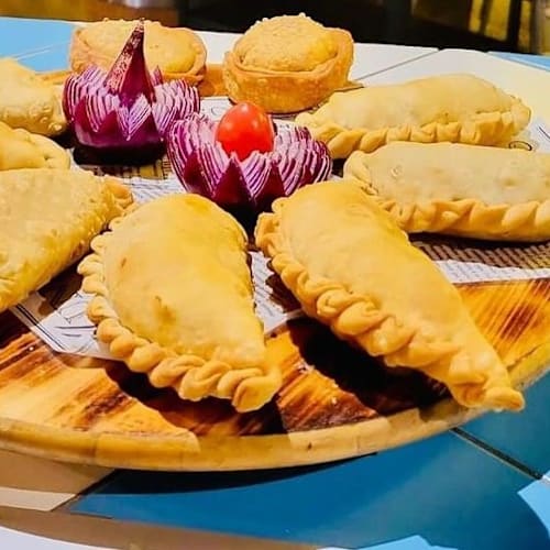 Empanadas doradas y crujientes, acompañadas de pasteles morados en forma de flor y dulces redondos.