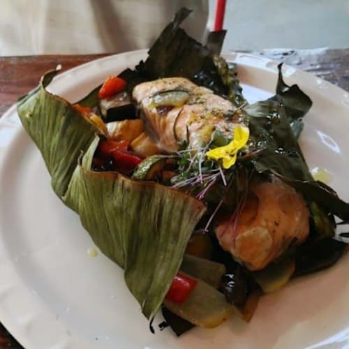 Salmón envuelto en hojas de plátano, acompañado de verduras asadas y decorado con microgreens y una flor comestible.