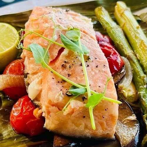 Filete de salmón envuelto en hojas de plátano, acompañado de tomates cherry asados, espárragos y una rodaja de lima.