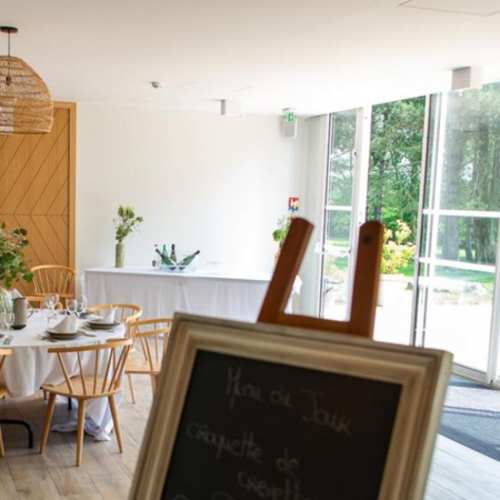 Intérieur de restaurant moderne avec une table ronde dressée, nappée de blanc, ornée d'un vase de verdure, et baignée de lumière naturelle grâce à de grandes fenêtres.