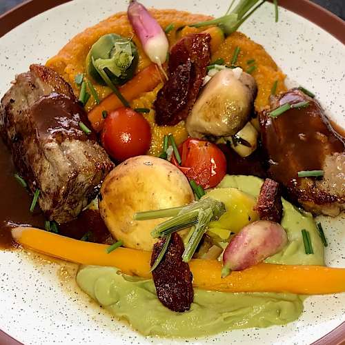 Plat de viande tendre, probablement rôti ou grillé, nappé d'une sauce riche, accompagné de légumes rôtis colorés et d'une purée verte crémeuse, le tout agrémenté d'herbes fraîches et de fleurs comestibles.