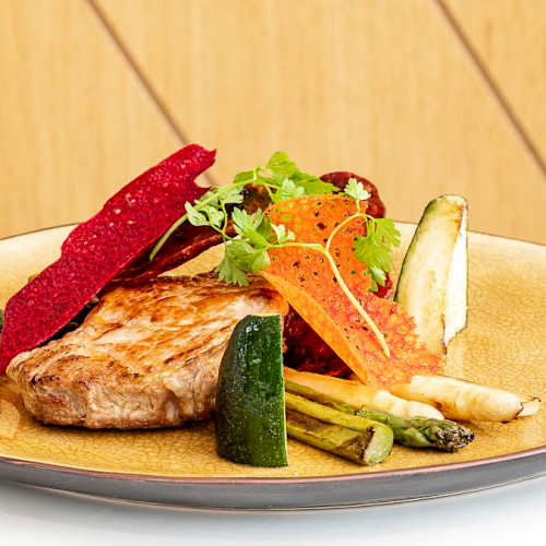 Plat de viande parfaitement grillée, accompagné d'un assortiment de légumes colorés et de chips de légumes croquants, le tout rehaussé d'herbes fraîches.