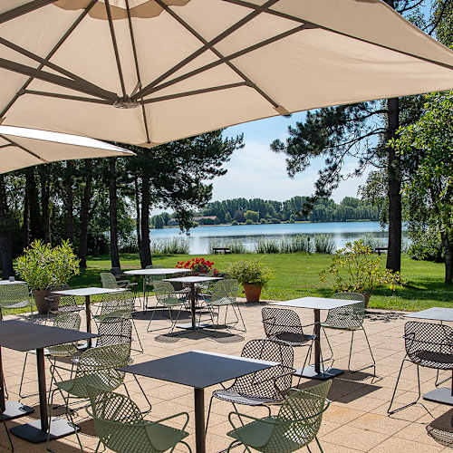 Espace de restauration en plein air avec des tables sur un patio carrelé, des chaises modernes, des parasols, et une vue sereine sur un lac entouré de verdure.