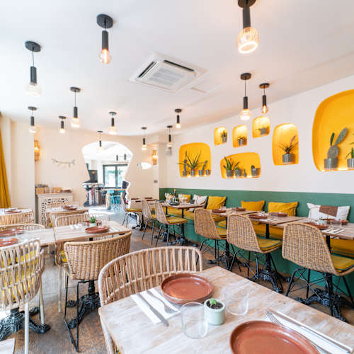 Int&eacute;rieur de restaurant lumineux et accueillant, alliant des &eacute;l&eacute;ments modernes et rustiques, avec de grandes fen&ecirc;tres, des rideaux jaunes, des chaises tiss&eacute;es autour de tables en marbre, et un mur d'accent vert. Des niches d&eacute;coratives mettent en valeur des plantes en pot, tandis que l'&eacute;clairage par ampoules suspendues cr&eacute;e une ambiance chaleureuse et contemporaine.
