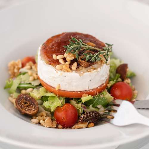 Queijo de cabra cremoso sobre uma cama de folhas verdes frescas, acompanhado de tomates-cereja e pinhões. O queijo é levemente caramelizado, com fatias de figos secos que acrescentam textura e sabor. Um toque de tomilho enriquece a apresentação do prato.