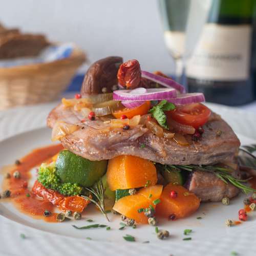 Um prato bem apresentado com um corte espesso de carne, provavelmente porco, sobre uma cama colorida de legumes. A carne é guarnecida com fatias de cebola roxa, ervas frescas e tomates-cereja, proporcionando um toque de cor. À volta da carne, encontram-se pedaços tenros de courgette, cenouras laranja e pimentos grelhados, todos regados com um molho rico que realça a apresentação. O prato demonstra um equilíbrio de texturas e sabores, prometendo uma experiência gastronómica satisfatória.