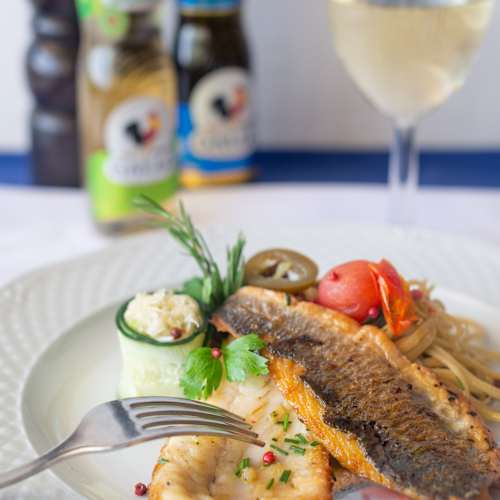 Prato elegante com dois tipos de peixe, um com pele crocante e o outro tenro e esfarelado, guarnecido com ervas frescas e acompanhado de um molho cremoso, tomates-cereja e uma fatia de pepino. Acompanhado por massa ligeiramente temperada.