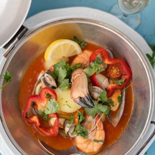 Uma mistura de marisco com camarões, amêijoas e mexilhões, servida num caldo rico à base de tomate, guarnecida com coentros frescos e fatias de pimento vermelho, acompanhada de gomos de limão.