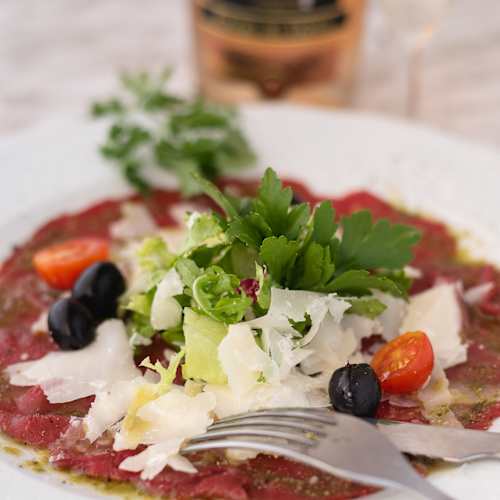 Carpaccio de carne de vaca com fatias finas de carne crua, regado com um molho leve e coberto com uma mistura fresca de alface e salsa. Tomates-cereja e azeitonas pretas adicionam cor e sabor, enquanto lascas de queijo parmesão enriquecem o prato com um toque salgado.