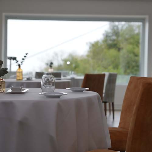 Interior moderno de restaurante con mesa redonda, manteles blancos, sillas de color beige y grandes ventanales que aportan luz natural y vistas al exterior.